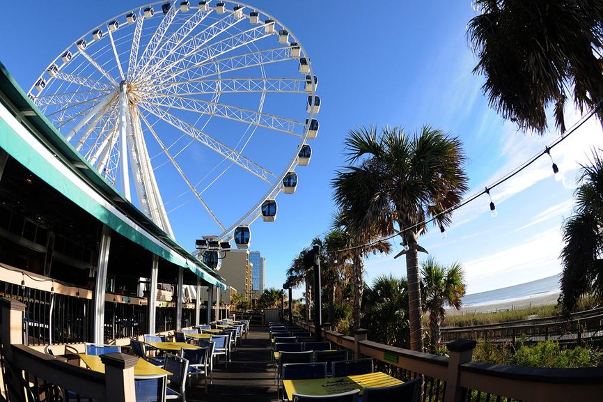 Oceanfront deck with restaurant on left side, palm trees and beach on the right and ferris wheel in the background