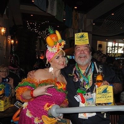 Woman in a colorful tropical dress with a fruit hat posing next to a man wearing a paper party hat