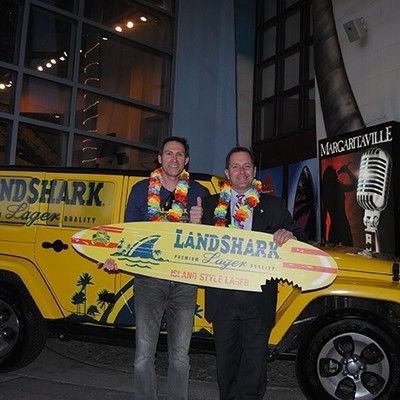 Two men holding a LandShark surfboard sign in front of a branded yellow vehicle