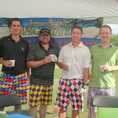 Four men in golf attire and patterned Loudmouth shorts standing under a event tent