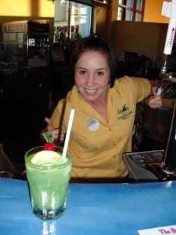 Smiling female bartender in a yellow shirt posing behind a green tropical drink