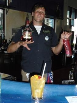 Male bartender holding liquor bottles behind a colorful tropical drink at the bar