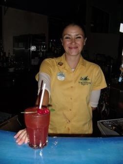 Smiling female bartender in a yellow shirt standing behind a red tropical cocktail