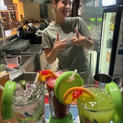 Photo of female bartender showing thumbs up behind an assortment of colorful cocktails