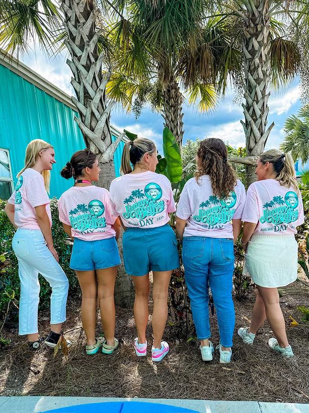 Women with their backs to the camera to show off their Jimmy Buffett day t=shirts that have the logo on the back