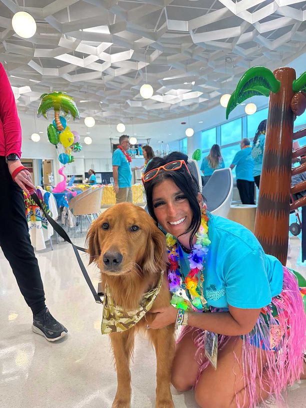 A woman smiling, wearing a grass skirt and lei necklace kneeling next to a golden retriever