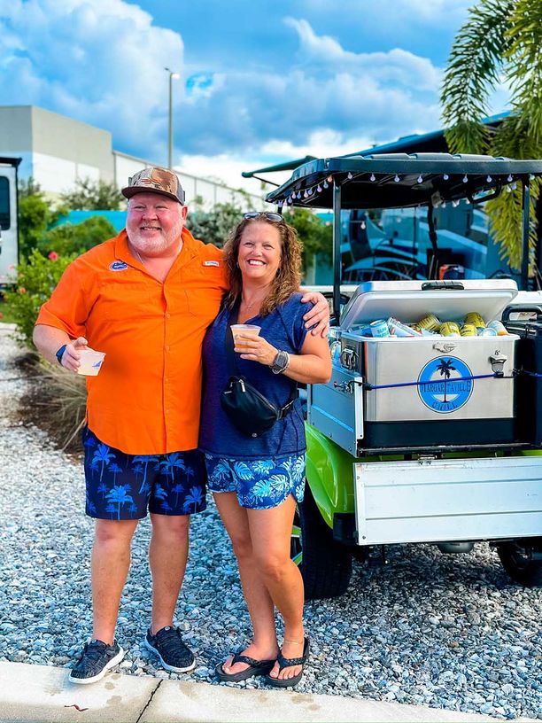 Man and woman smiling while holding drinks next to a golf cart with an ice chest in the back full of drinks