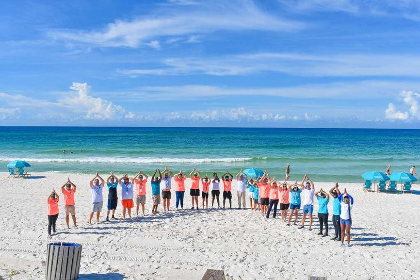 Large group of people ina a half circle on the beach doing the Fins Up sign with their hands