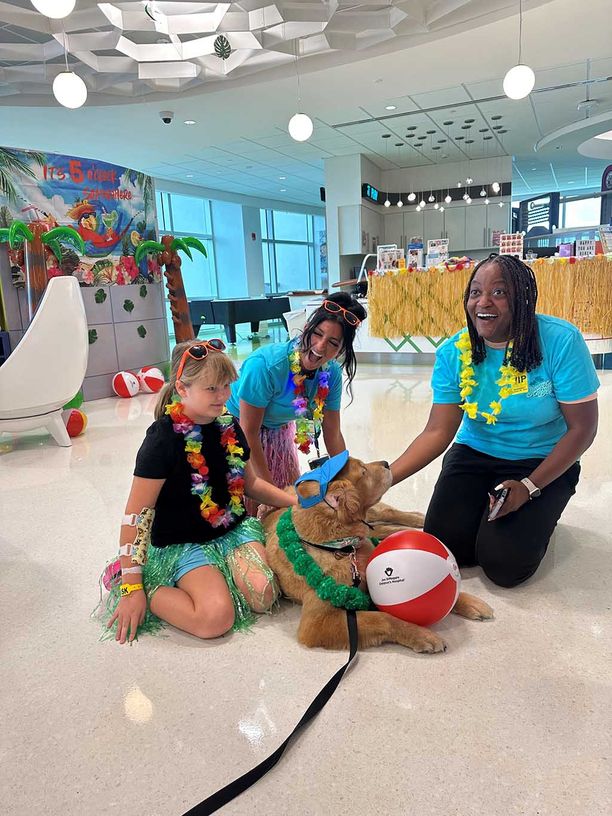 Two women and a girl in lei necklaces playing with a goldn retriever dog 