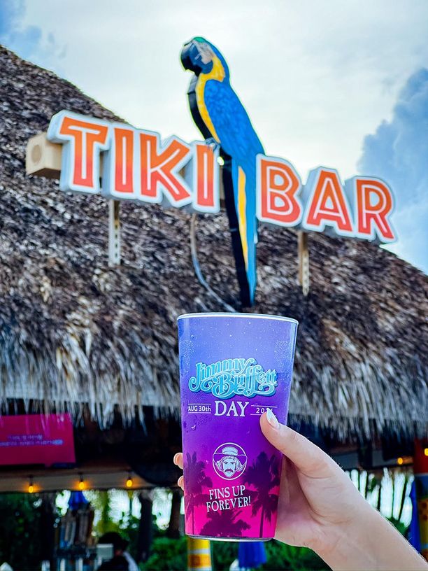 A Jimmy Buffett Day cup being held in front of a Tiki Bar sign