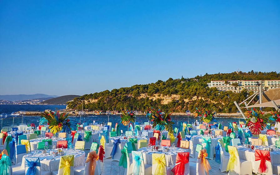 Tables with white linen clothes and multicolored ribbons on the chairs grouped outside with a hill and a beautiful blue body of water in the background