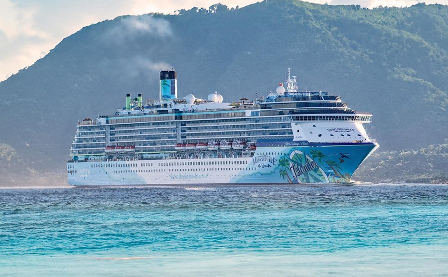 Margaritaville at Sea Islander cruise ship docked near a lush green island under a bright, clear sky, surrounded by tranquil turquoise waters.