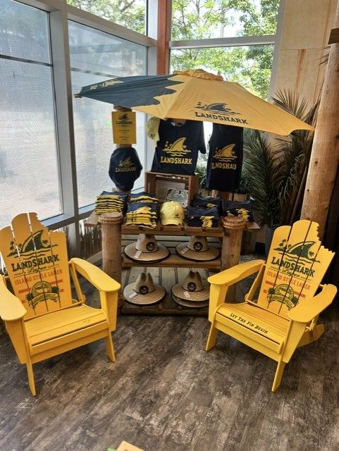 LandShark Lager merchandise display with yellow umbrella, dark blue shirts, straw hats, and two yellow beach chairs