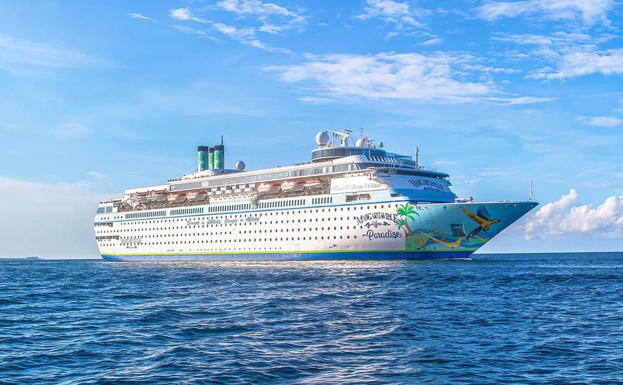 Margaritaville at Sea Paradise cruise ship sailing on a clear blue ocean under a sunny sky, with a backdrop of clouds and horizon.