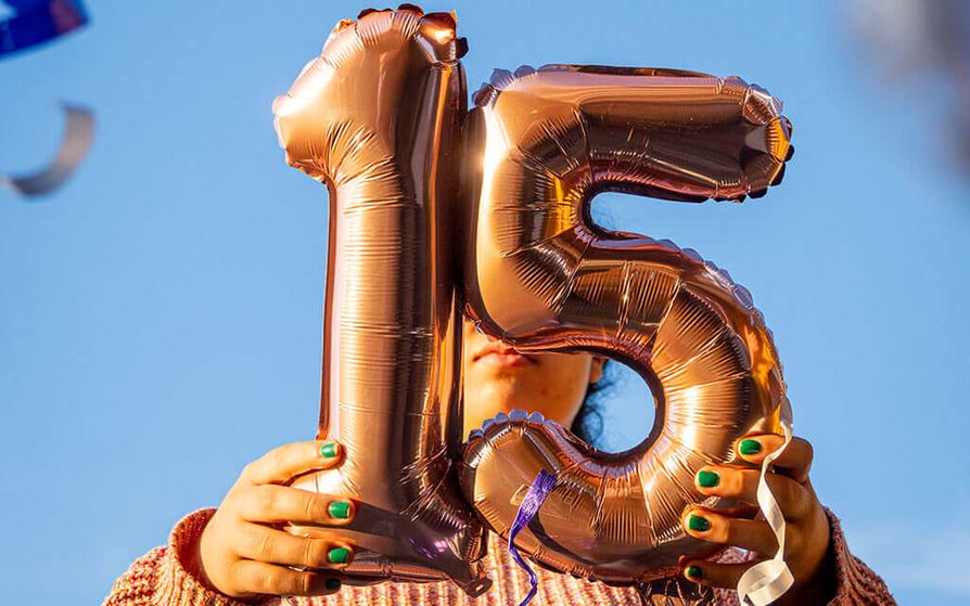 Person with green painted nails holding a golden balloon of the number 15 in front of their face