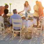 Three men and three women dine at a table on the beach